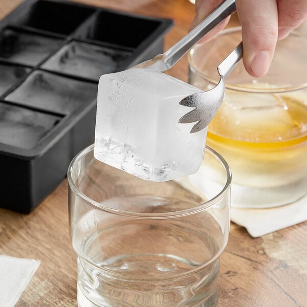 A hand uses metal tongs to place an ice cube in a glass.