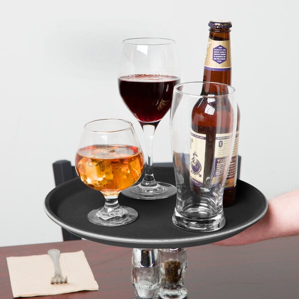 A person holding a Carlisle black non-skid serving tray with wine glasses and a bottle on it.