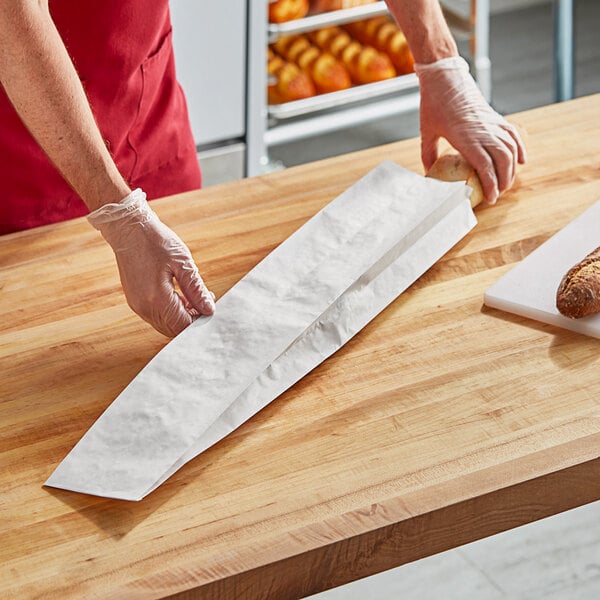 A person using a plastic glove to place a baguette in a Bagcraft bread bag on a counter.