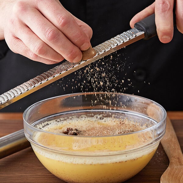 A person grating whole nutmeg over a bowl of yellow liquid.