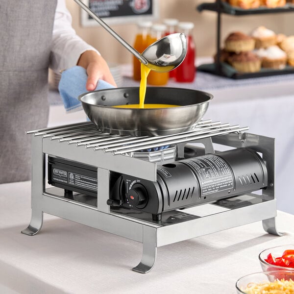 A silver steel butane burner frame with a grill top, shown heating a pan as liquid is poured in.
