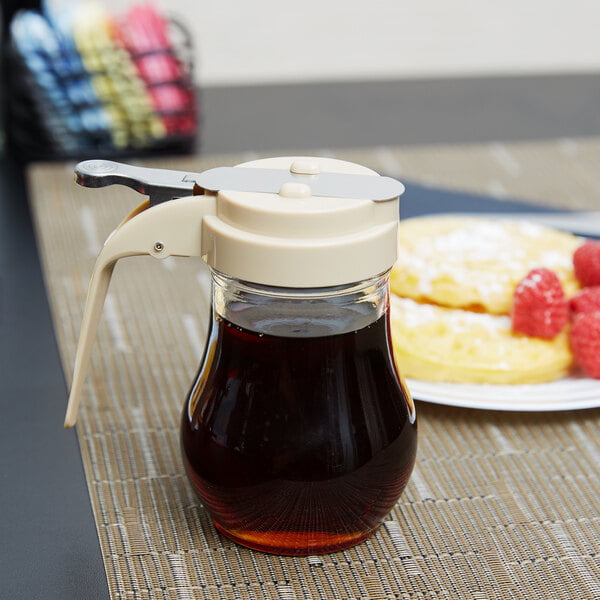 A Tablecraft glass syrup dispenser filled with brown syrup on a table with a spoon.