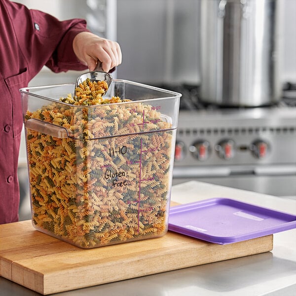 A woman pouring pasta into a Vigor clear food storage container on a school kitchen counter.