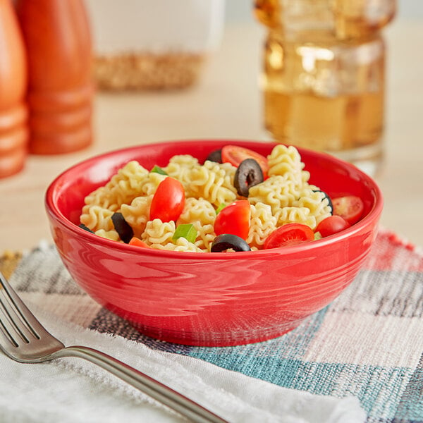 A close-up of an Acopa Capri Passion Fruit Red stoneware bowl filled with pasta, tomatoes, and olives on a cloth with a fork.