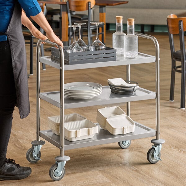 A three-shelf stainless steel utility cart holding glassware, bottles, plates, and trays in a restaurant setting.
