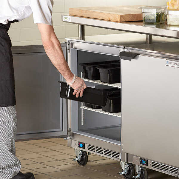 A person in a blue shirt and gloves putting a black tray into a Beverage-Air undercounter freezer.