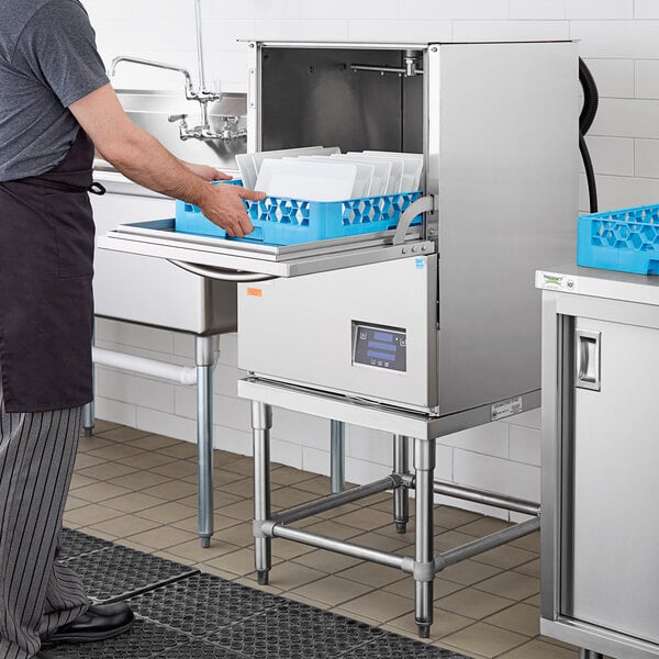 A man standing in a kitchen using a Noble Warewashing undercounter dishwasher with white plates in a blue basket.