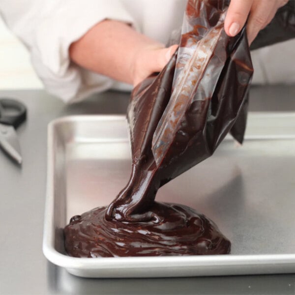 A person pouring chocolate brownie batter from a plastic bag onto a metal baking tray.
