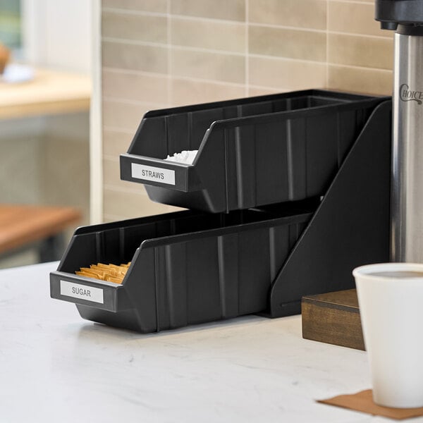 A black two-tier organizer with labeled bins for straws and sugar, placed on a countertop.