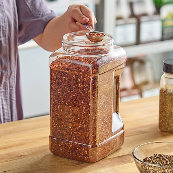A woman pouring red flakes into a 1 gallon square PET plastic jar on a table.