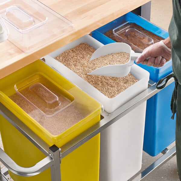 A person using a white scoop to pour grains into a Baker's Lane ingredient bin on a counter.