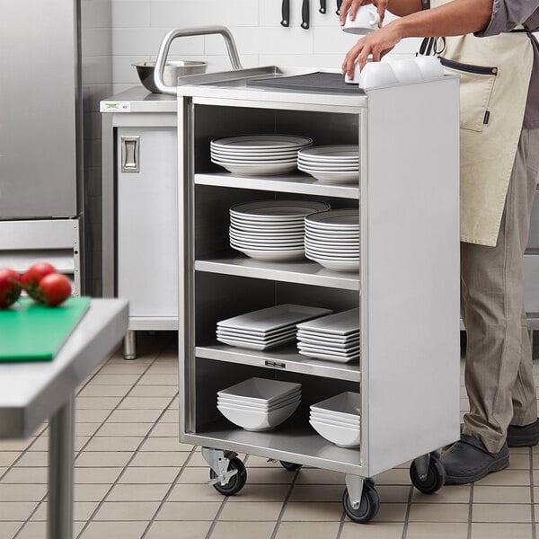 A man standing in a kitchen next to a Regency stainless steel utility cart with plates on the shelf.