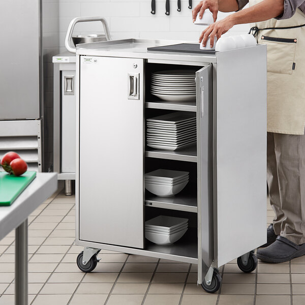 A person standing next to a Regency stainless steel utility cart with plates and bowls on it.