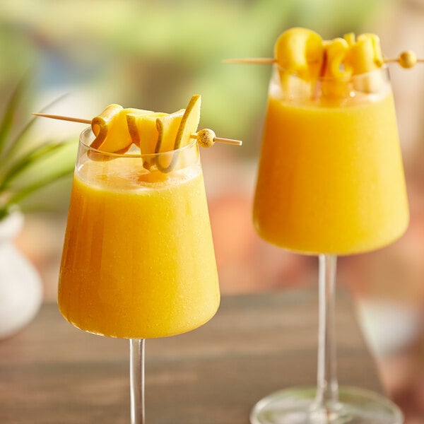 Two glasses of yellow liquid with fruit on a stick.