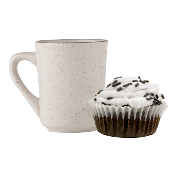 A speckled beige mug next to a chocolate cupcake with white frosting and chocolate sprinkles in a white fluted baking cup.