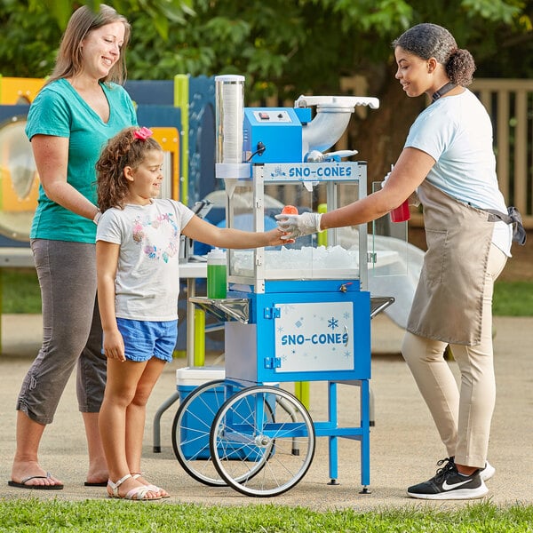 A woman and a girl standing next to a red and white Carnival King cart with a small blue cup of shaved ice.