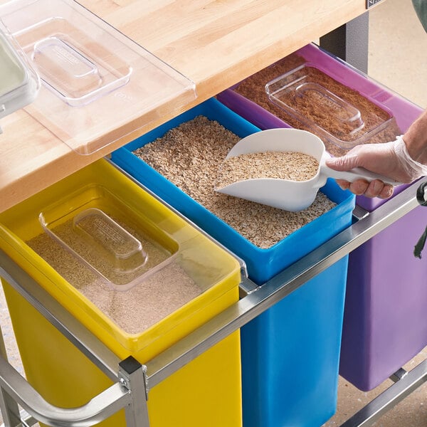 A person pouring grains into a yellow, blue, and purple Baker's Lane ingredient bin using a handle.