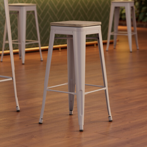 Three Lancaster Table & Seating white metal backless bar stools with gray wood seats.