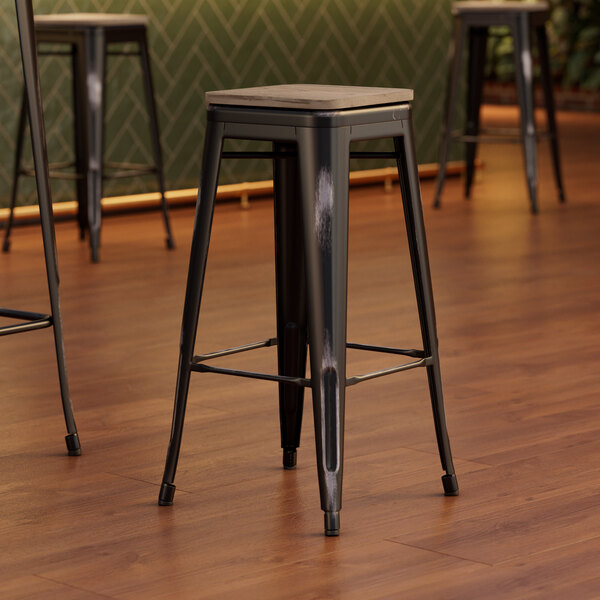 Three Lancaster Table & Seating black metal barstools with gray wood seats on a wood floor.