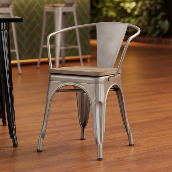 A Lancaster Table & Seating silver metal arm chair with a gray wood seat on a wood floor.