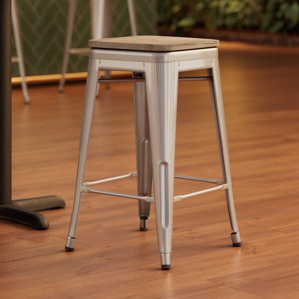 A Lancaster Table & Seating silver metal stool with a gray wood seat on a wood floor.