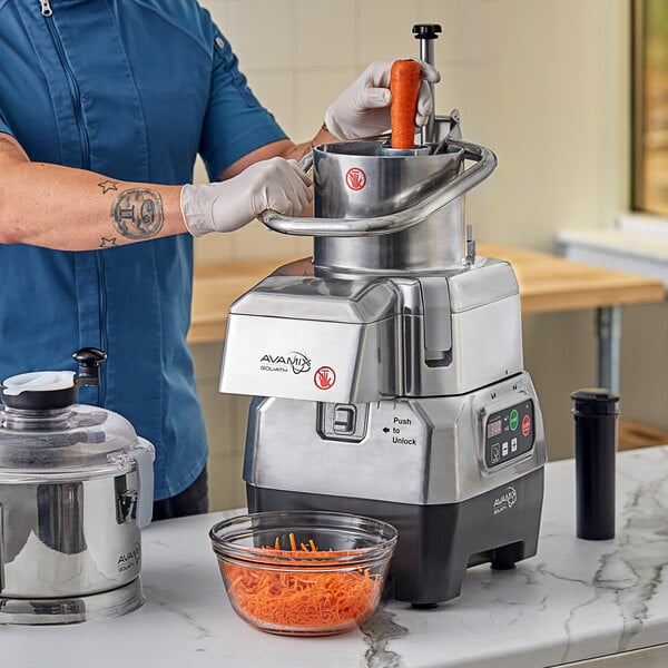 A man in a blue uniform and gloves using an AvaMix stainless steel bowl on a food processor to shred carrots.
