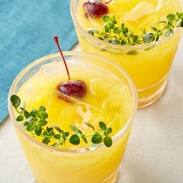 Two glasses of yellow liquid with cherries and herbs on a table in a cocktail bar.