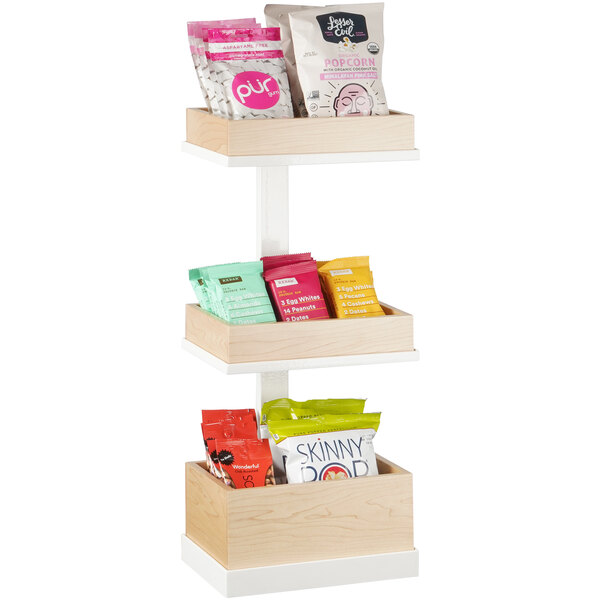 A Cal-Mil wooden three tier merchandiser with snacks on it including popcorn, cotton candy, and a red package.
