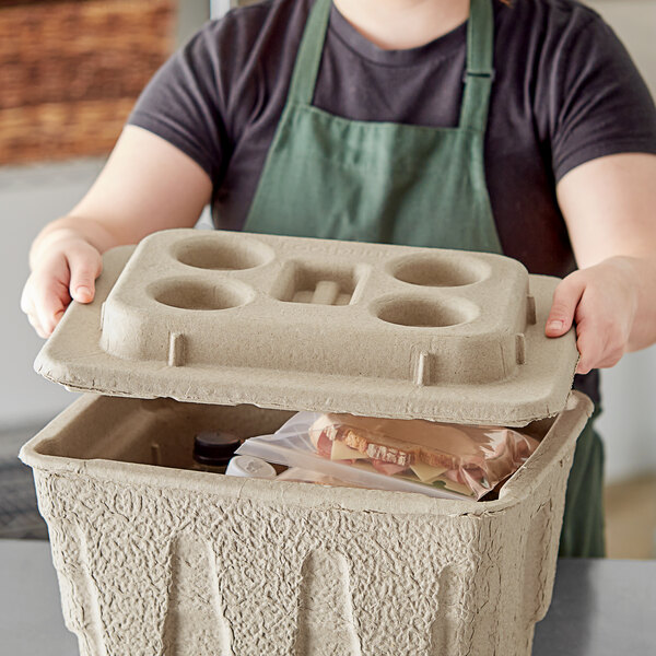A woman wearing a green apron holding a Fiber Cooler Lid on a container of food.