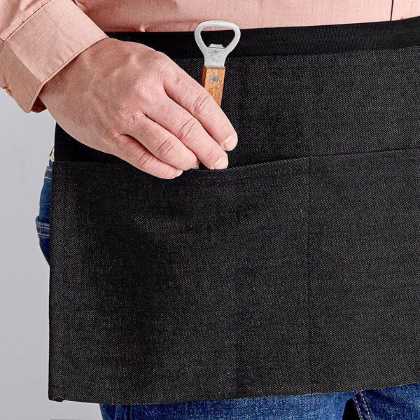 A person holding a bottle opener in the pocket of a black Acopa Kennett waist apron.