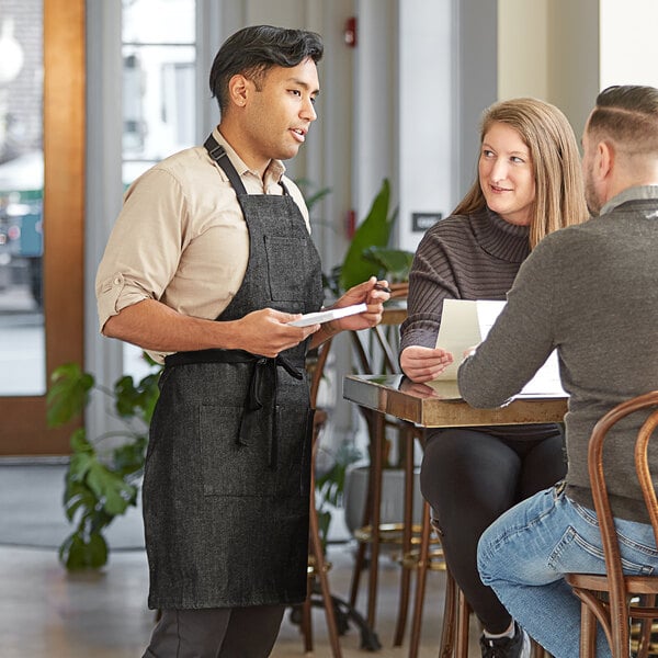 A man in a black Acopa Kennett apron standing next to a woman sitting at a table.