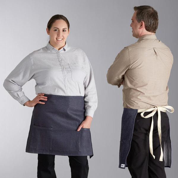 A man and woman standing next to each other, both wearing blue denim half bistro aprons with natural webbing.
