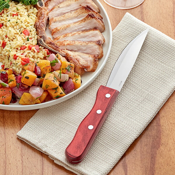 An Acopa steak knife with a cherry finish next to a plate of meat and rice with vegetables.