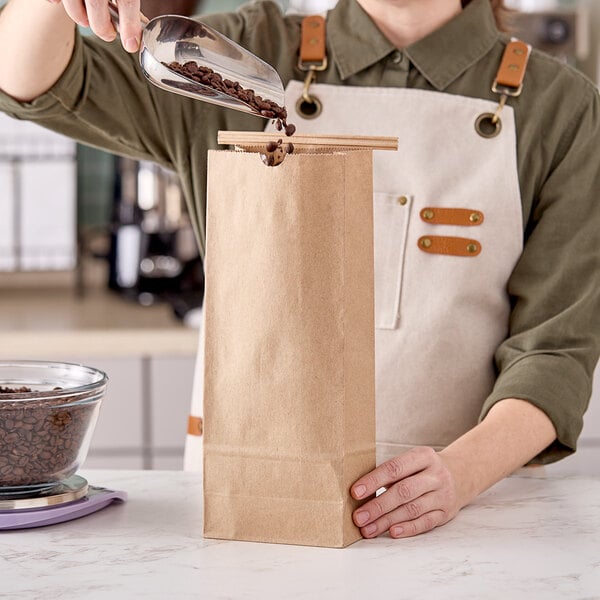 A person using a metal scoop to pour coffee beans into a brown paper bag.