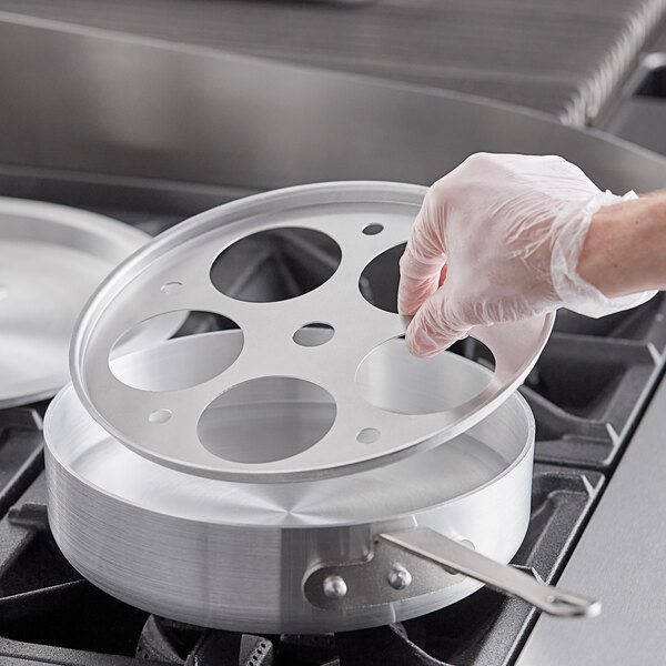 A person in a plastic glove holding a Choice aluminum egg poacher inset over a pan.