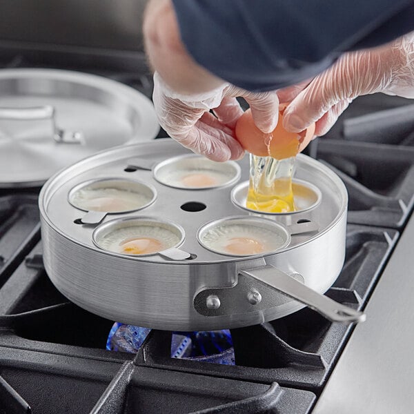 A person breaking an egg into a pan with Choice 5-Cup Egg Poacher Set on a stove.