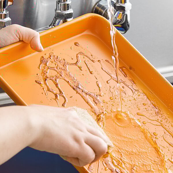 A person washing a Baker's Mark orange non-stick sheet tray on a counter with a sponge.