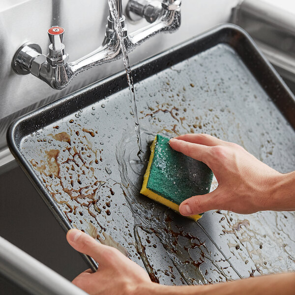 A hand cleaning a Baker's Mark sheet pan on a counter with a sponge.