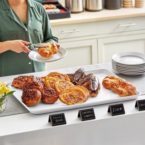 A woman holding a Baker's Mark white wire rim aluminum sheet tray of pastries on a table.