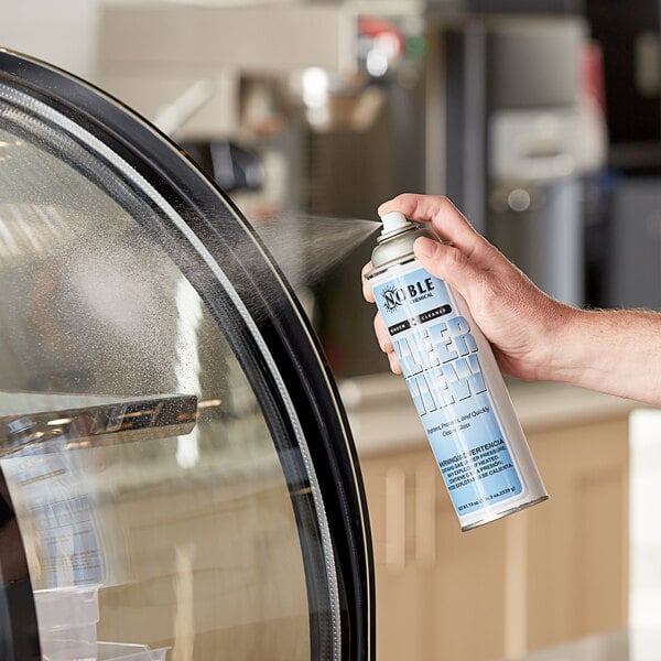 A hand holding a can of Noble Chemical Kleer View glass cleaner spraying a glass door.