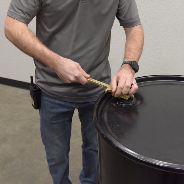A person using a non-sparking bronze alloy drum bung nut wrench to open a large black drum with a 2 3/4 inch opening.