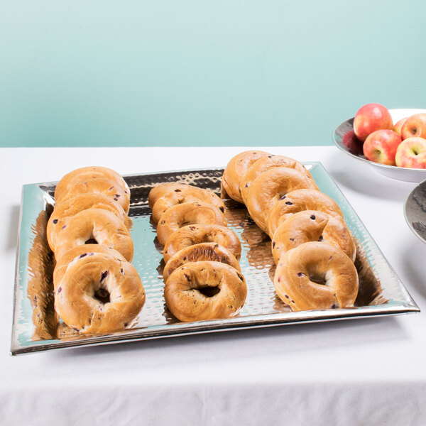 A close up of a tray of bagels and a bowl of apples on a hammered stainless steel tray.