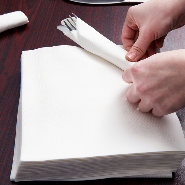 A person folding a white Hoffmaster FashnPoint dinner napkin with a fork and knife inside.