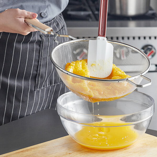 A person using a Vollrath medium mesh strainer with a wood handle to strain eggs.