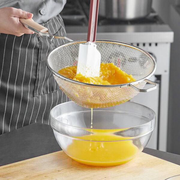 A person stirring egg yolk in a bowl with a Vollrath medium mesh wire strainer with a wood handle.