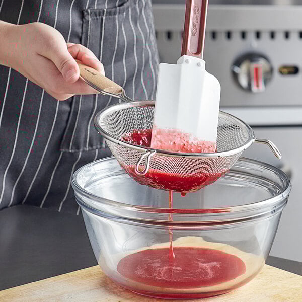 A person using a Vollrath medium mesh wire strainer with a wood handle to pour red liquid into a bowl.