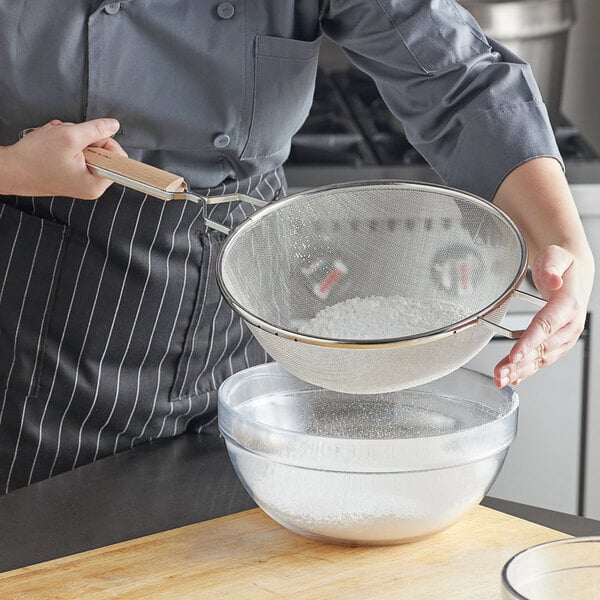 A woman using a Vollrath fine mesh strainer with a wood handle to sift white powder into a bowl.