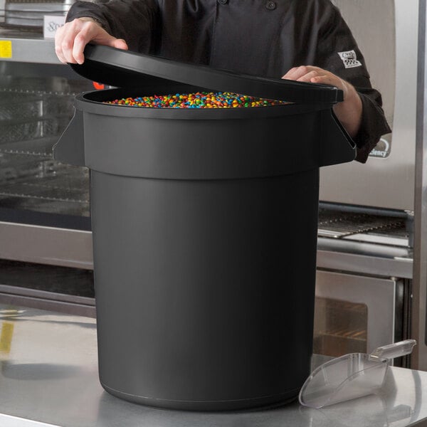 A man in a chef's uniform opening a large black round ingredient storage bin filled with colorful candies.