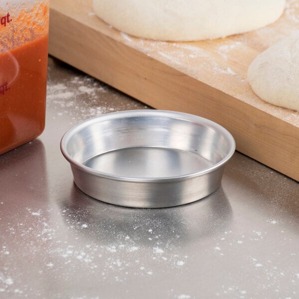 A metal bowl with dough in it on a white counter next to a round silver American Metalcraft pizza pan.