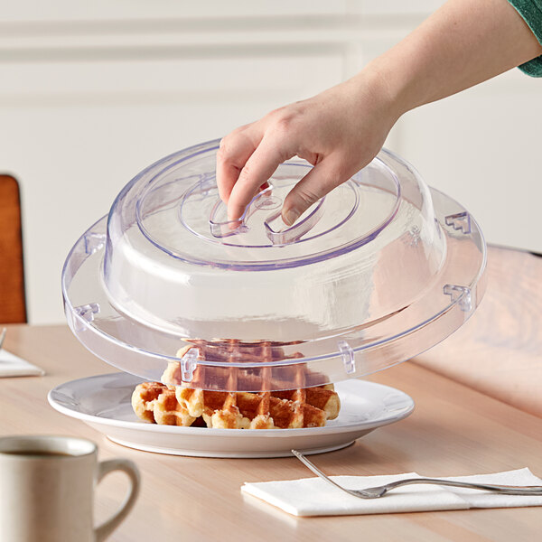 A person using an American Metalcraft clear plastic plate cover to place a lid on a plate of waffles on a table.